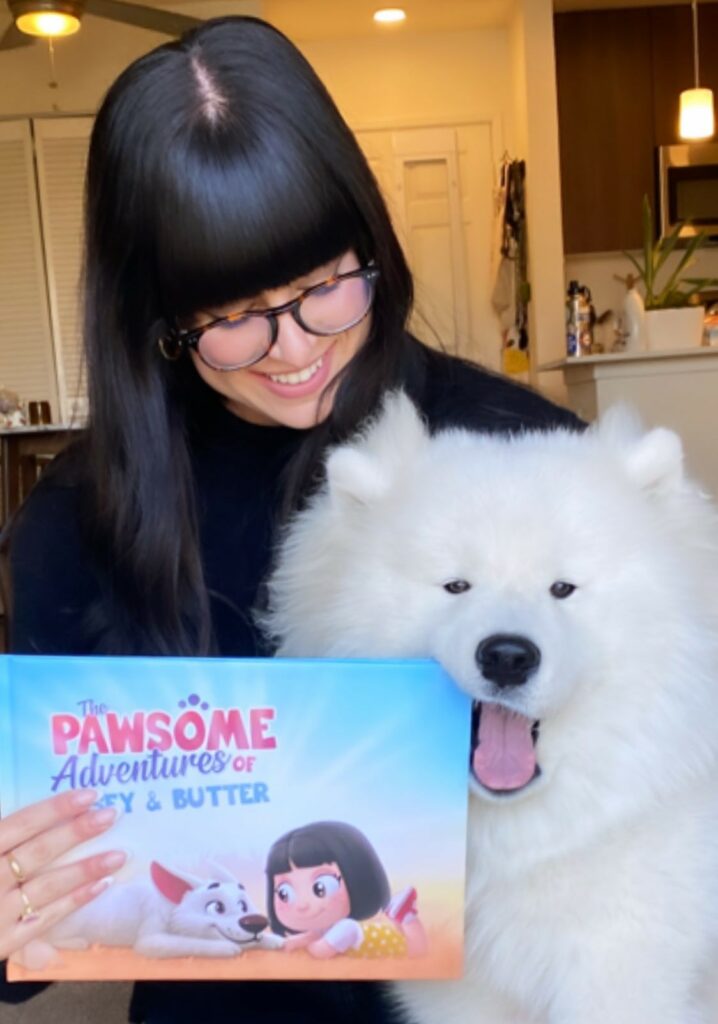 A woman cuddles a white dog who is chewing on the corner of a custom book for pets and their humans.