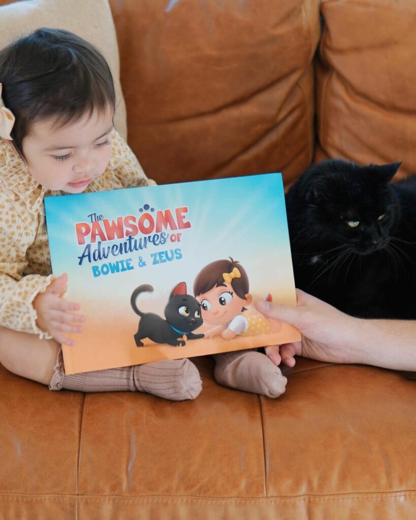 A little girl sits with her cat on the floor and their custom book for her and her cat.