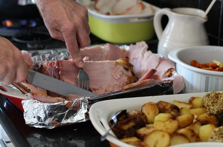 A man slices up a Christmas ham next to a tray of roast potatoes and veggies. 