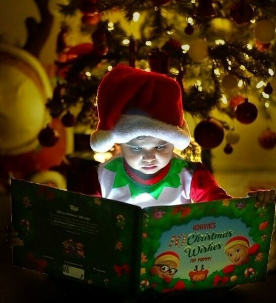 A child dressed as an elf reads his own personalized Christmas book for Mommy and child in front of an xmas tree.