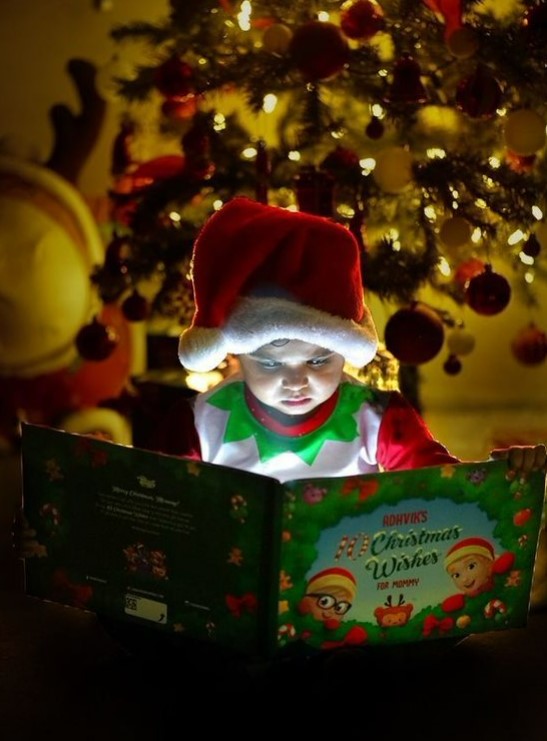 A child dressed as an elf reads his own personalized Christmas book for Mommy and child in front of an xmas tree.