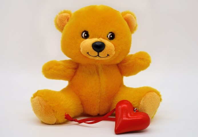 An orange teddy bear sitting in front of a red heart ornament.