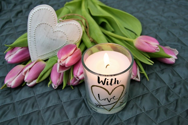 A valentine's day candle, pink tulips and a white heart ornament on a table.
